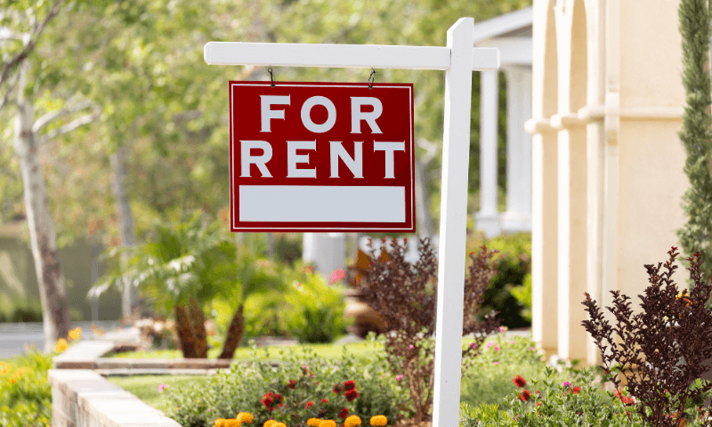 A red sign reading "for rent" hangs on a white holder in the front yard of a house surrounded by trees and flowers.
