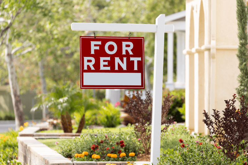 A red sign reading "for rent" hangs on a white holder in the front yard of a house surrounded by trees and flowers.