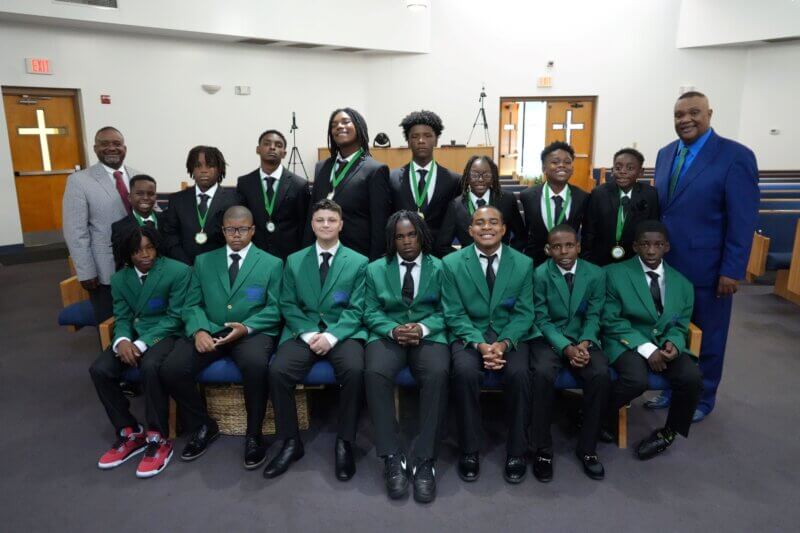 One line of boys sits on a church pew while another line stands behind them.